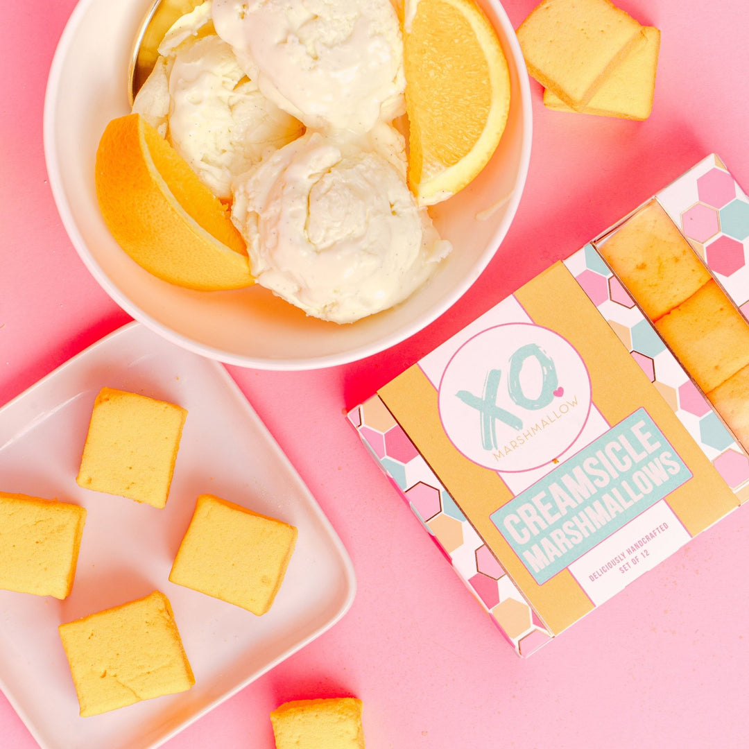 A box of creamsicle marshmallows against a pink background next to a bowl of vanilla ice cream with orange slices in it