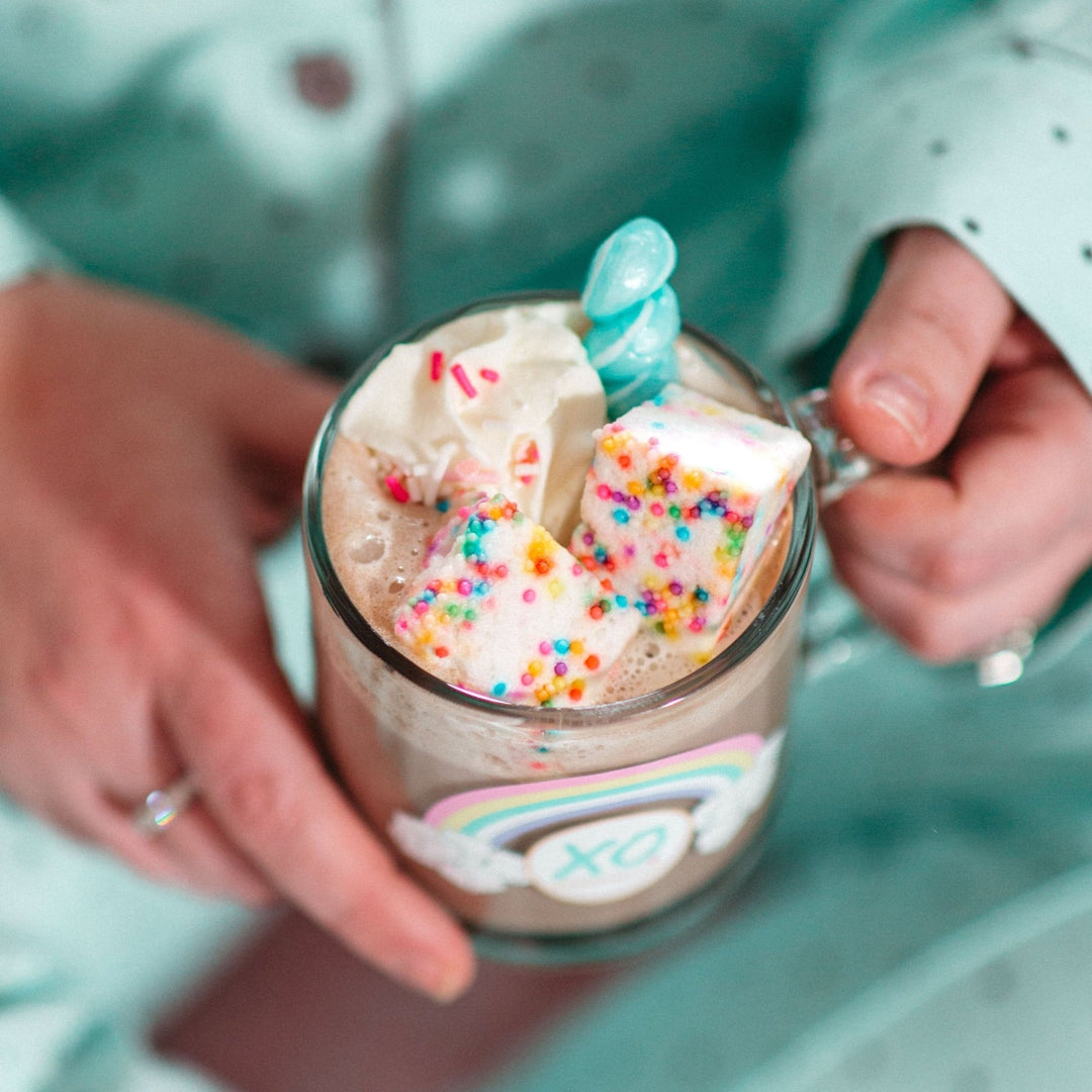 A person in mint polka dot pajamas holding a glass mug full of hot cocoa topped with XO Marshmallow Funfetti Gourmet Marshmallows