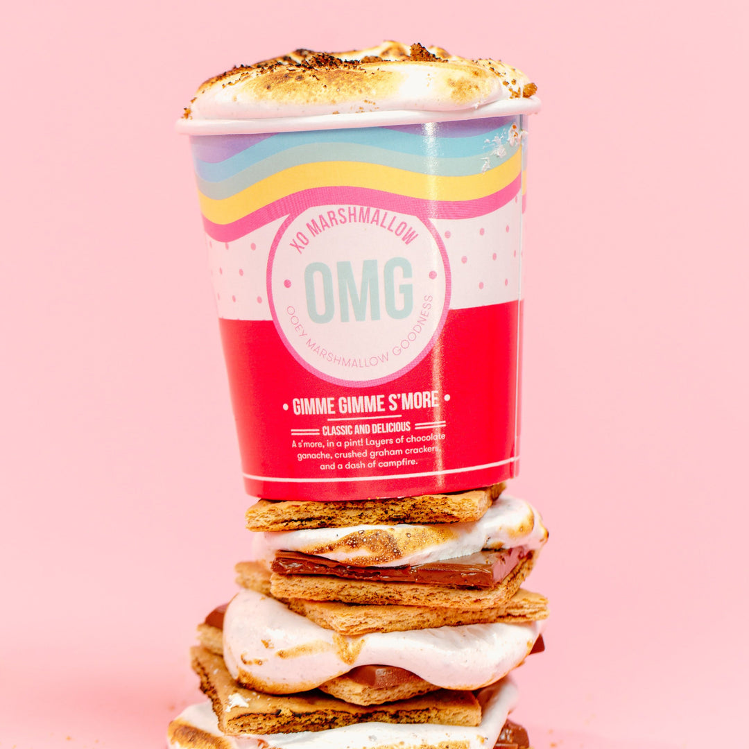 A pint container of marshmallow creme on top of s'mores with layers of chocolate ganache and crushed graham crackers, with a decorative presentation on a pink background.
