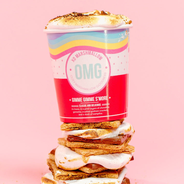 A pint container of marshmallow creme on top of s'mores with layers of chocolate ganache and crushed graham crackers, with a decorative presentation on a pink background.