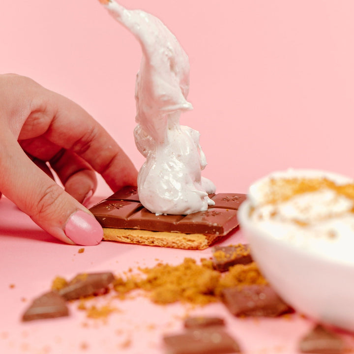 A hand holding the base of a s'more which is a graham cracker and bar of chocolate while it scoops gimmie s'more marshmallow on top of the chocolate with a spoon