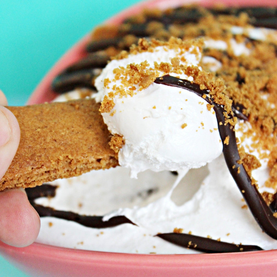 A bowl of gimmie s'more gourmet marshmallow spreadable creme topped with chocolate ganache and crushed gluten free graham crackers. A hand is using another graham cracker to dip it into the fluff
