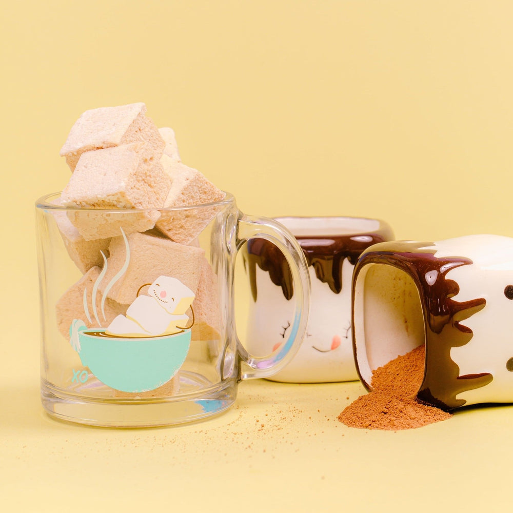 A glass cup featuring a marshmallow snowman filled with a stack of gourmet XO Marshmallow hot chocolate flavored marshmallows. There are 2 more mugs in the photo to the right and the mugs are shaped like marshmallows with faces. One of them is knocked over and hot cocoa mix is spilling out of it. All of this is against a yellow background.