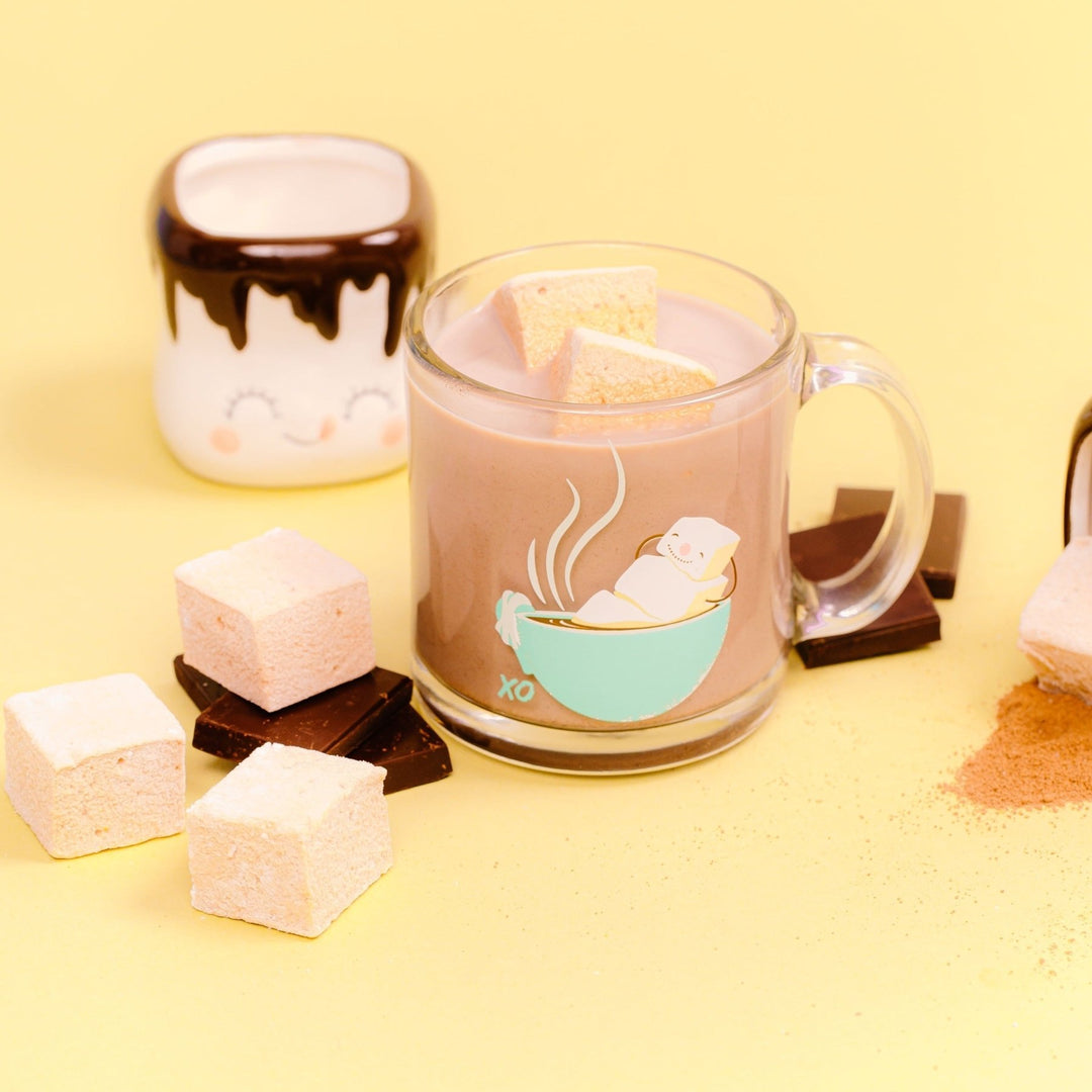 A glass coffee mug filled with hot chocolate and 2 melted hot chocolate flavored marshmallows is in the center. There is a small mug shaped like a marshmallow with a face in the left background. On both sides of the glass mug are pieces of chocolate bars with hot chocolate flavored marshmallows on top of them. All of this is against a yellow background.