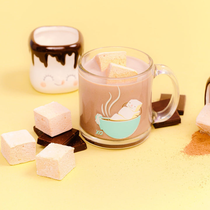 A glass coffee mug filled with hot chocolate and 2 melted hot chocolate flavored marshmallows is in the center. There is a small mug shaped like a marshmallow with a face in the left background. On both sides of the glass mug are pieces of chocolate bars with hot chocolate flavored marshmallows on top of them. All of this is against a yellow background.