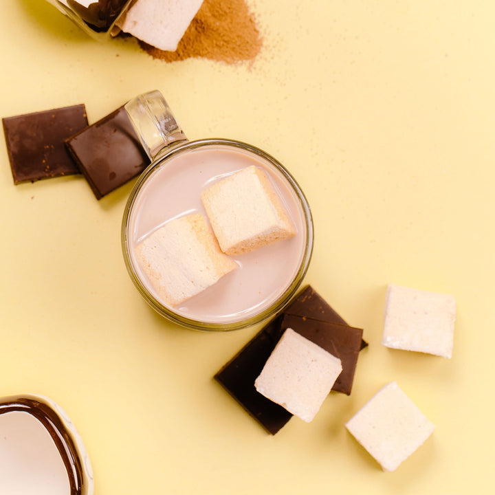 A glass cup filled with hot chocolate, surrounded by white chocolate pieces and chocolate-flavored marshmallows on a yellow background.