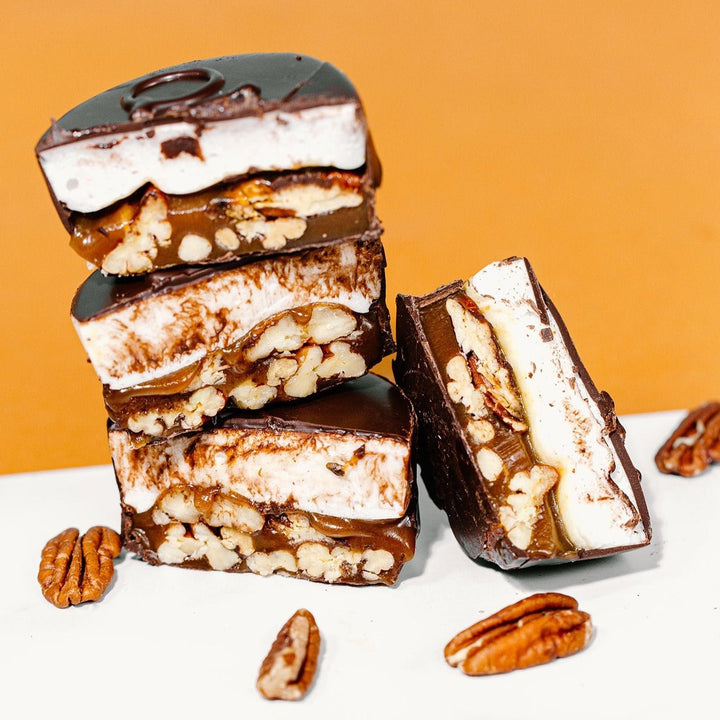 Stack of marshmallow turtles with layers of chocolate, caramel, marshmallow and pecans visible, on a white surface with pecans scattered around.