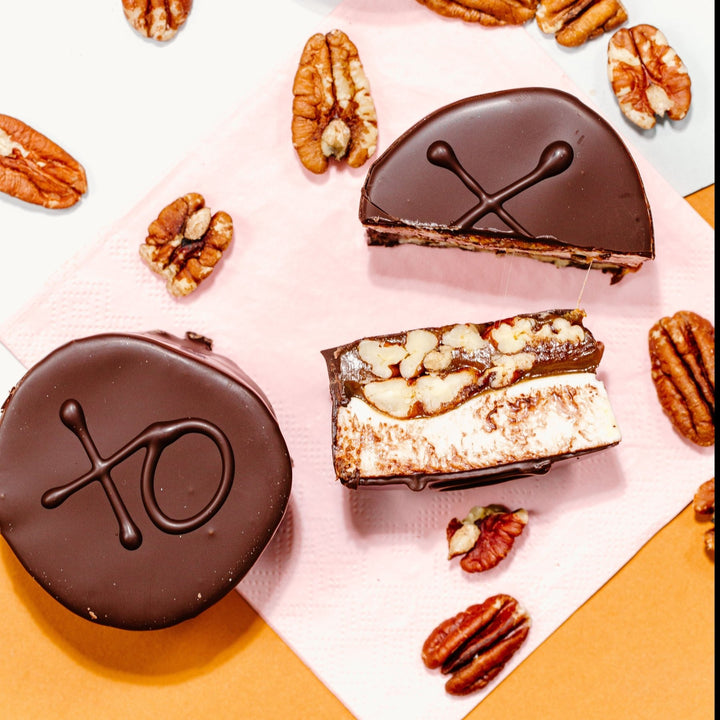 A marshmallow turtle candy fully coated in chocolate with an XO on top of it written in chocolate next to a turtle that's been cut in  half so you can see the caramel, pecans, and marshmallow inside.