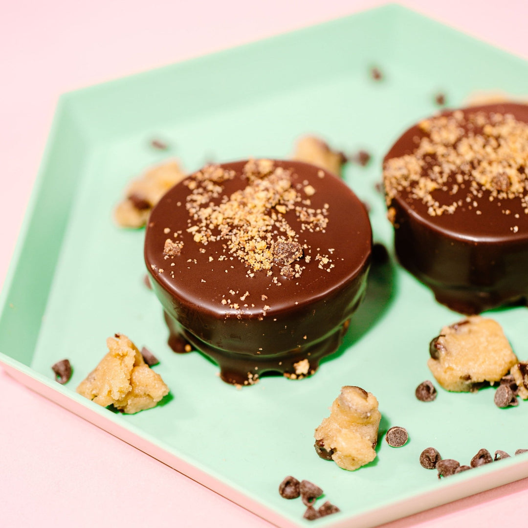 Two round Oh Dough You Didn't Cookie Dough cups against a hexagon mint plate. The cups are covered in chocolate and topped with crushed cookies 