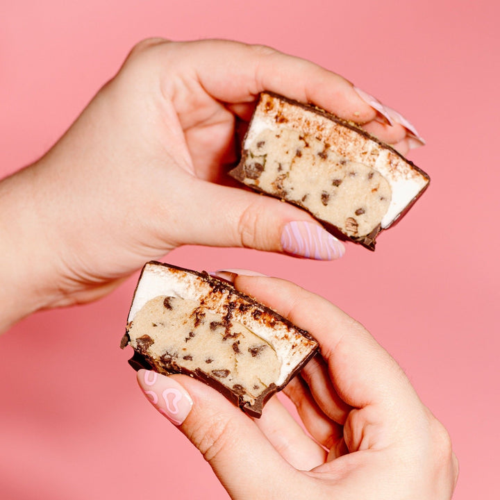 Two hands each holding half of a Oh Dough You Didn't Gluten Free Edible Chocolate Chip Cookie Cup showing the cookie dough and marshmallow inside. This is against a pink background