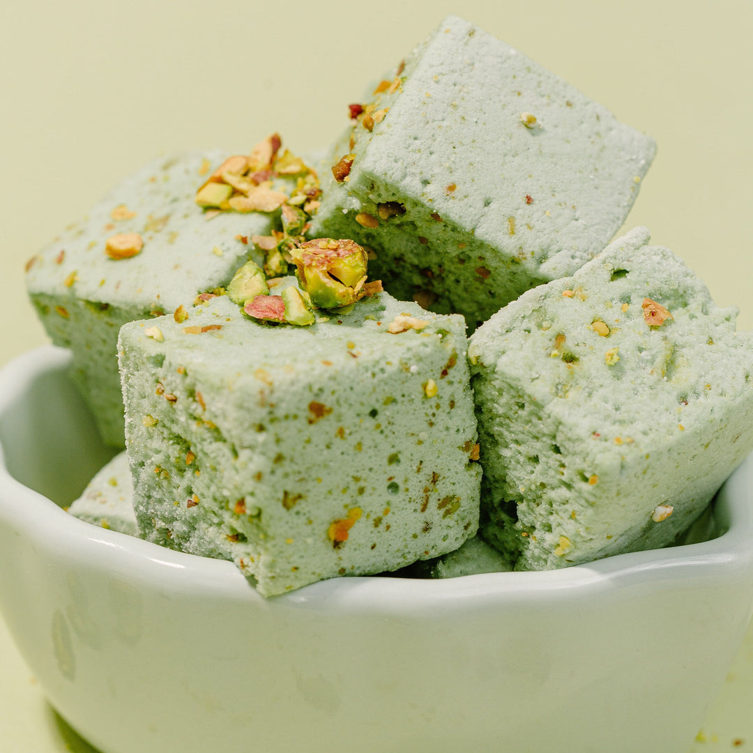 XO Marshmallow Gourmet Pistachio marshmallows stacked in a pile in a light green bowl against a green background. Crushed pistachios are drizzled on top of the marshmallows to demonstrate flavor.
