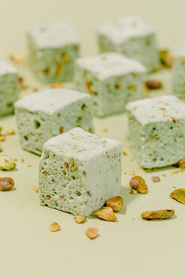 Eight Pistachio marshmallows are on their side in a grid formation against a light green background. You can see chunks of chopped pistachios inside of the marshmallows for added texture.