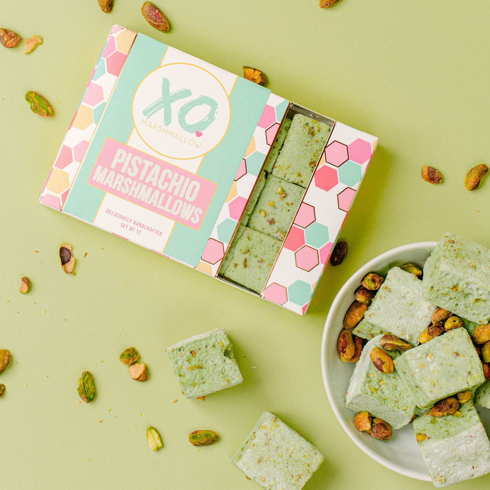 Pistachio Marshmallows inside of their packaging against a green background. The packaging is a colorful box with a window showing 3 pistachio marshmallows and the XO Marshmallow logo. Next to the box is a bowl of marshmallows and shelled pistachios are drizzled across the photo.