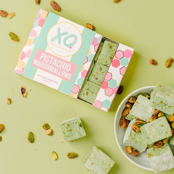Pistachio Marshmallows inside of their packaging against a green background. The packaging is a colorful box with a window showing 3 pistachio marshmallows and the XO Marshmallow logo. Next to the box is a bowl of marshmallows and shelled pistachios are drizzled across the photo.