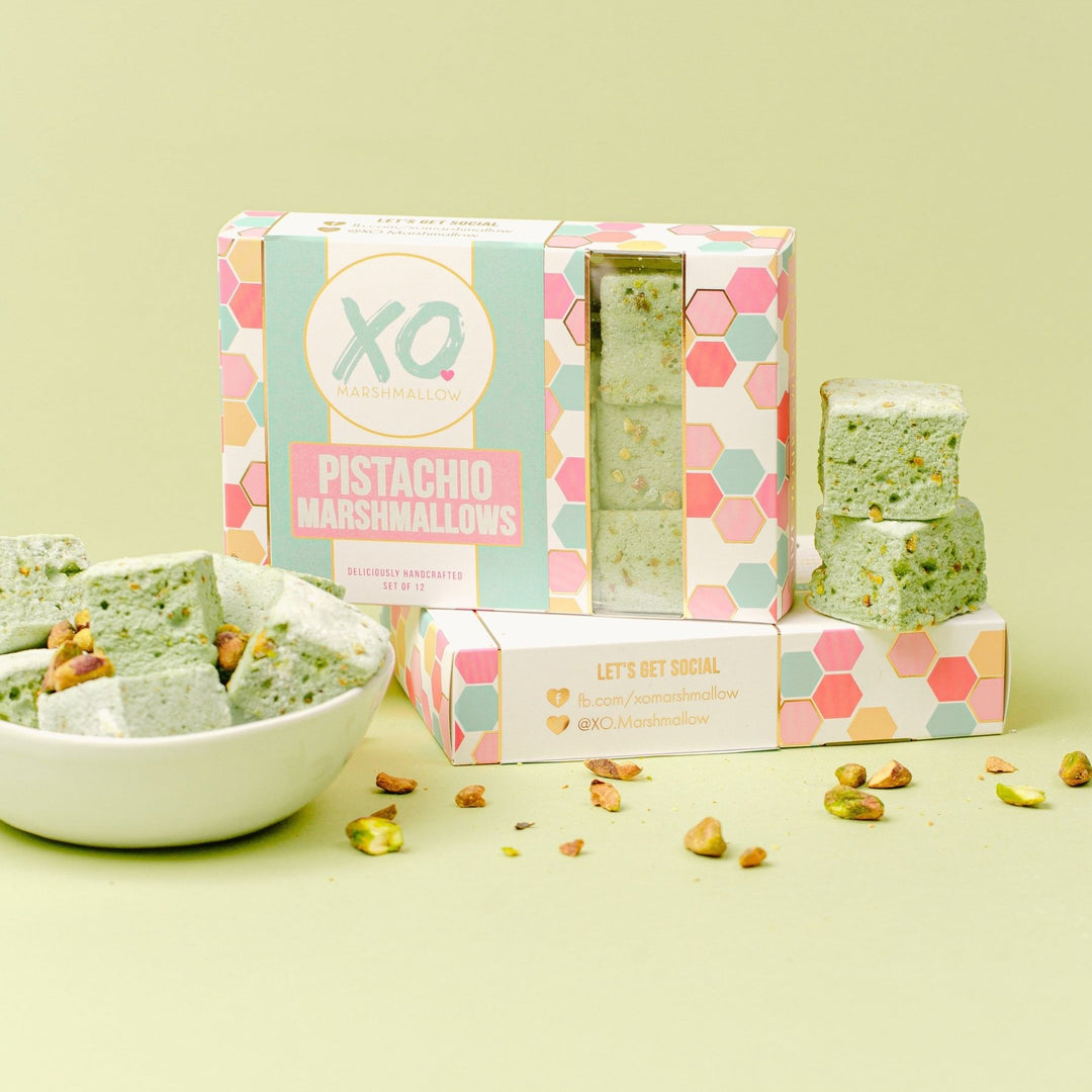 A Box of XO Marshmallow pistachio marshmallows standing up inside of their packaging on top of another box of packaging that's on it's side. Next to the box are two pistachio marshmallows and to the left of the box is a bowl full of pistachio marshmallows and pistachios.