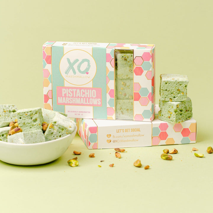 A Box of XO Marshmallow pistachio marshmallows standing up inside of their packaging on top of another box of packaging that's on it's side. Next to the box are two pistachio marshmallows and to the left of the box is a bowl full of pistachio marshmallows and pistachios.