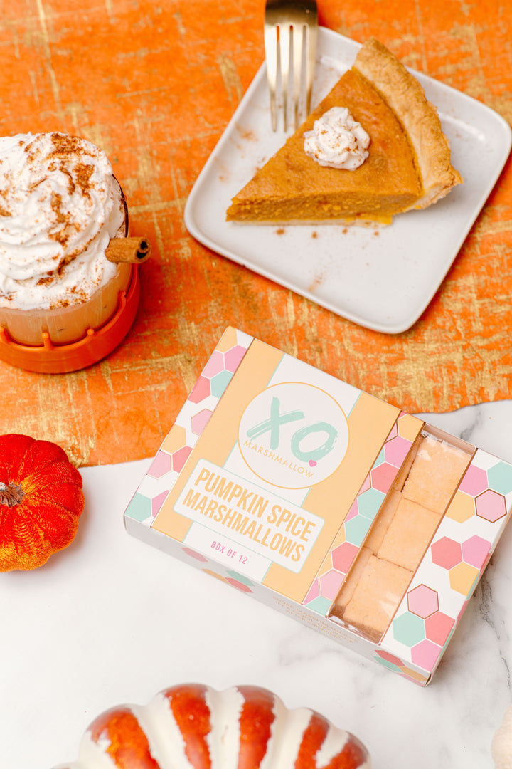 A package of Pumpkin Spice Marshmallows by XO is displayed on an orange textured background with decorative pumpkins and a slice of pumpkin pie, indicating the fall season and the product's pumpkin spice flavor.