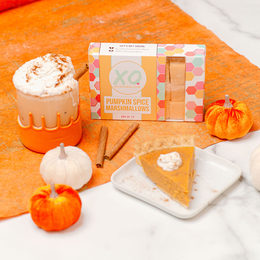 A package of Pumpkin Spice Marshmallows by XO is displayed on an orange textured background with decorative pumpkins and a slice of pumpkin pie, indicating the fall season and the product's pumpkin spice flavor.