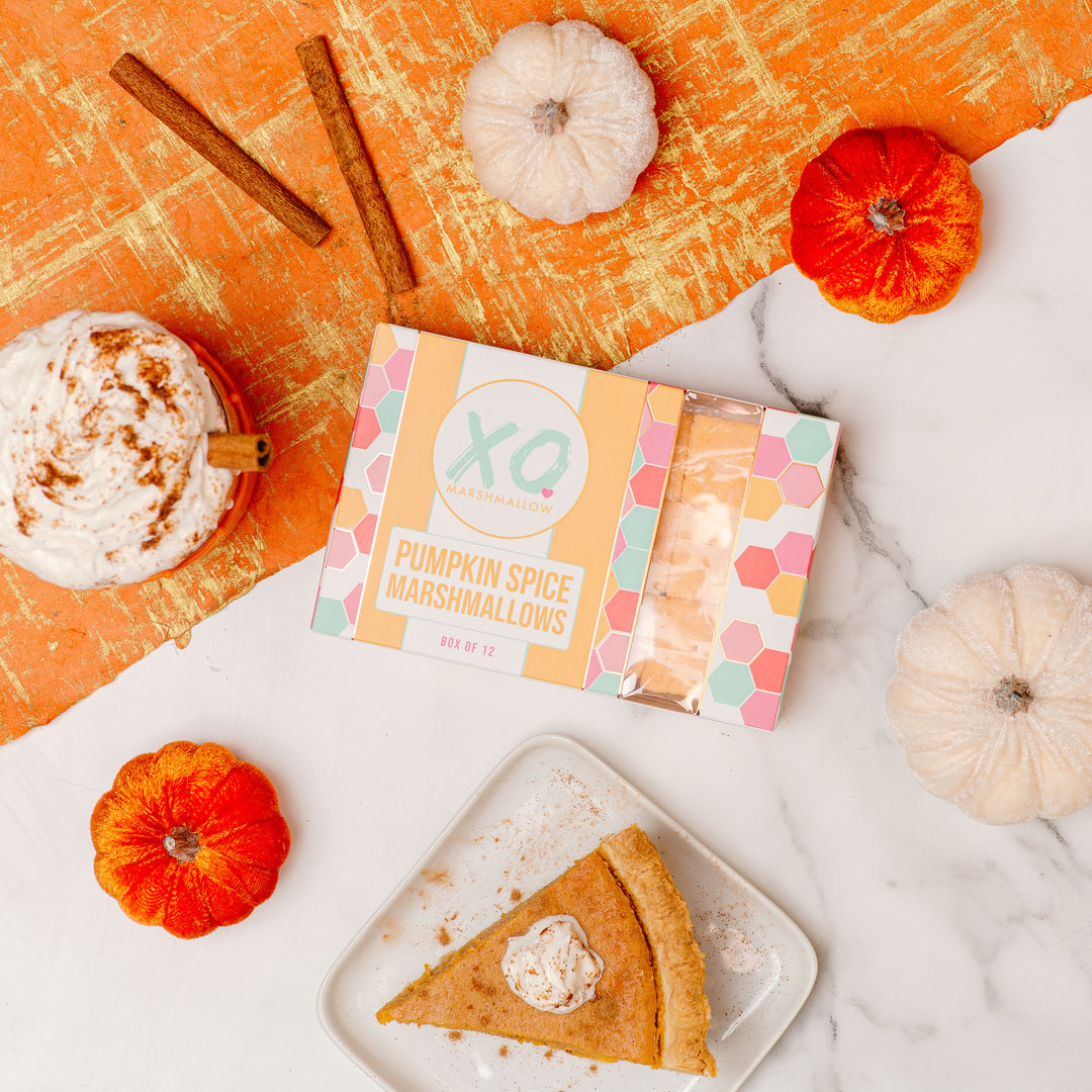 A package of Pumpkin Spice Marshmallows by XO is displayed on an orange textured background with decorative pumpkins and a slice of pumpkin pie, indicating the fall season and the product's pumpkin spice flavor.