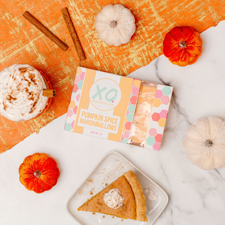 A package of Pumpkin Spice Marshmallows by XO is displayed on an orange textured background with decorative pumpkins and a slice of pumpkin pie, indicating the fall season and the product's pumpkin spice flavor.