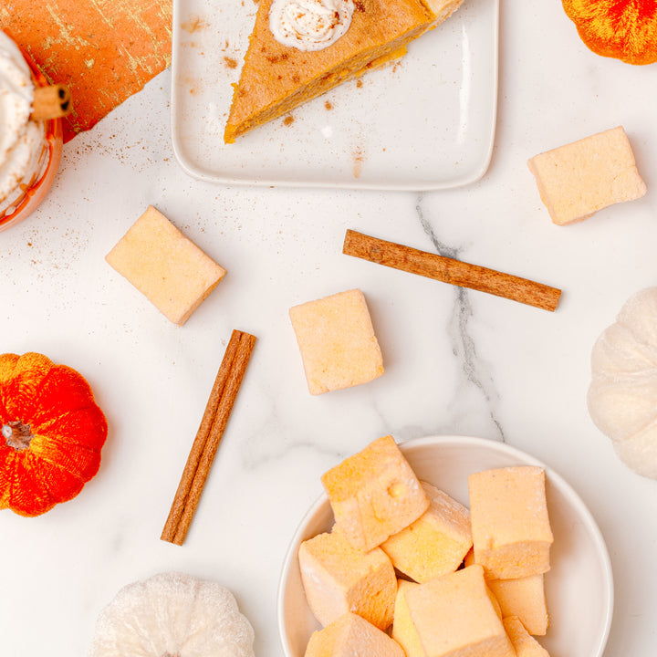 A package of Pumpkin Spice Marshmallows by XO is displayed on an orange textured background with decorative pumpkins and a slice of pumpkin pie, indicating the fall season and the product's pumpkin spice flavor.