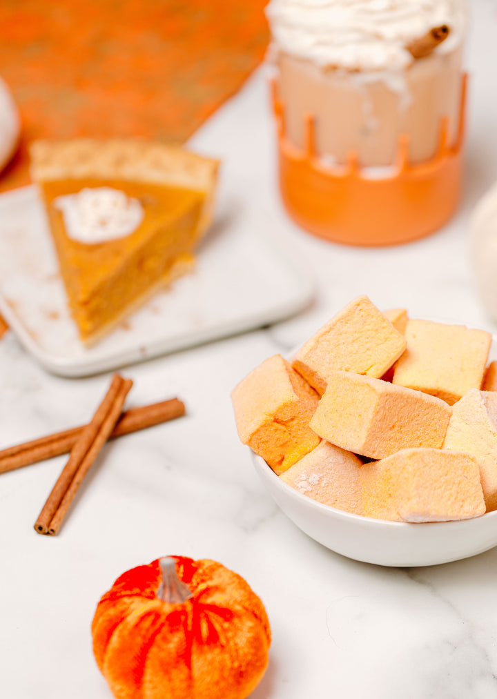 A package of Pumpkin Spice Marshmallows by XO is displayed on an orange textured background with decorative pumpkins and a slice of pumpkin pie, indicating the fall season and the product's pumpkin spice flavor.