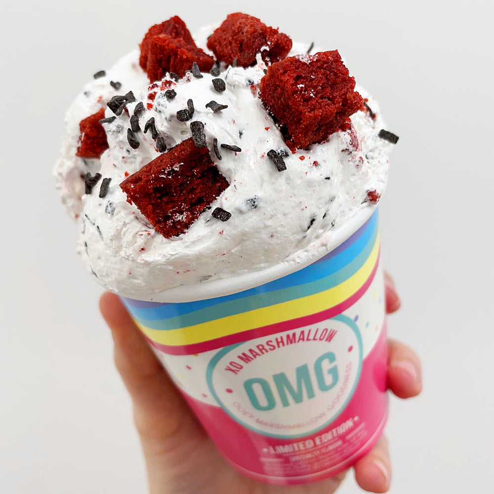 A person holding a pint of red velvet flavored marshmallow fluff featuring large chunks of gluten free red velvet cake and chocolate chip sprinkles on top against a white background. 