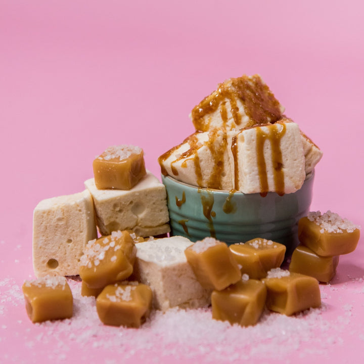 A bowl of homemade salted caramel marshmallows with caramel sauce drizzled over them and caramel candy pieces and rock salt sprinkled throughout the photo.