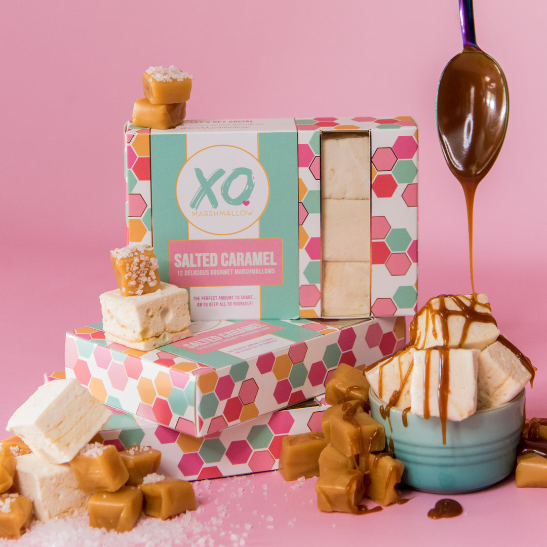A box of salted caramel marshmallows with some marshmallows displayed outside the box, alongside caramel pieces and a bowl of caramel sauce.