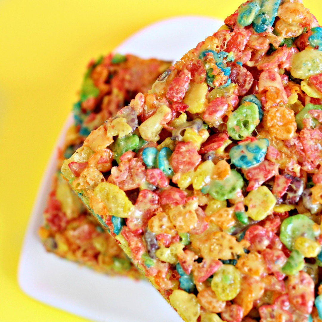 A colorful rainbow sherbet marshmallow dream bar with a layer of fruity pebbles crispy cereal on top, placed on a white plate against a yellow background.