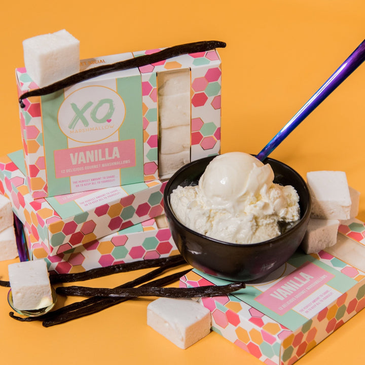 Boxes of XO Marshmallow vanilla marshmallows stacked up in their packaging. On top of one box is a bowl of vanilla ice cream with a spoon. Vanilla beans and vanilla marshmallows are scattered about the photo. Everything is against an orange background.