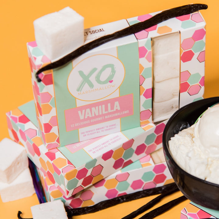 3 Boxes of XO Marshmallow vanilla marshmallows stacked up in their packaging. Peaking out of the bottom corner of the photo is a bowl of vanilla ice cream. Vanilla beans and vanilla marshmallows are scattered about the photo. Everything is against an orange background.