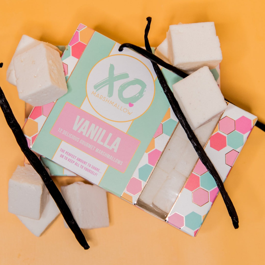 A set of vanilla marshmallows from XO Marshmallow, displayed alongside their packaging which prominently features the brand name and the flavor 'VANILLA'. The marshmallows are white and there are visible vanilla beans throughout.