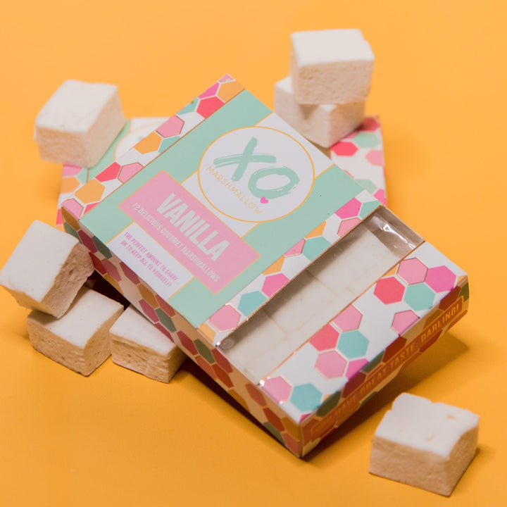 One box of XO Marshmallow Gourmet Vanilla Marshmallows laying flat against an orange background. Vanilla marshmallows are scattered around it.
