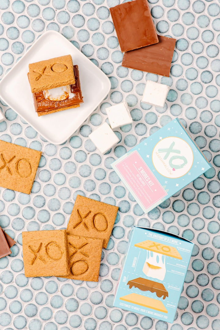 S'mores, marshmallows, graham crackers and chocolate bars are laid out against a blue penny tile background next to the blue box packaging that says XO Marshmallow Gourmet S'mores Kit. The side of the kit shows an illustration of a s'more being made