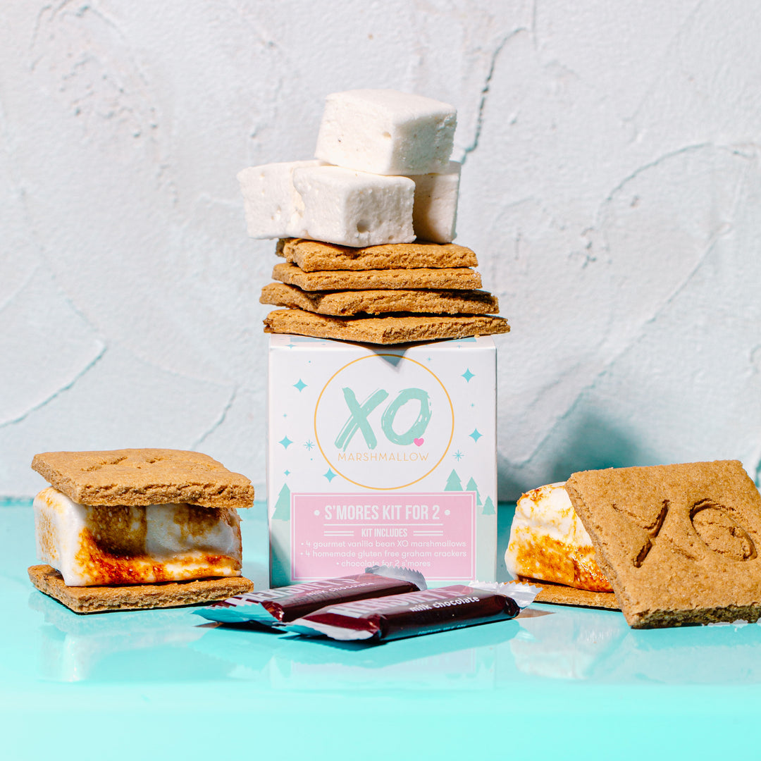 A s'mores kit for two people displayed with marshmallows, chocolate bars, and graham crackers arranged on a surface, with packaging labeled 'XO'.